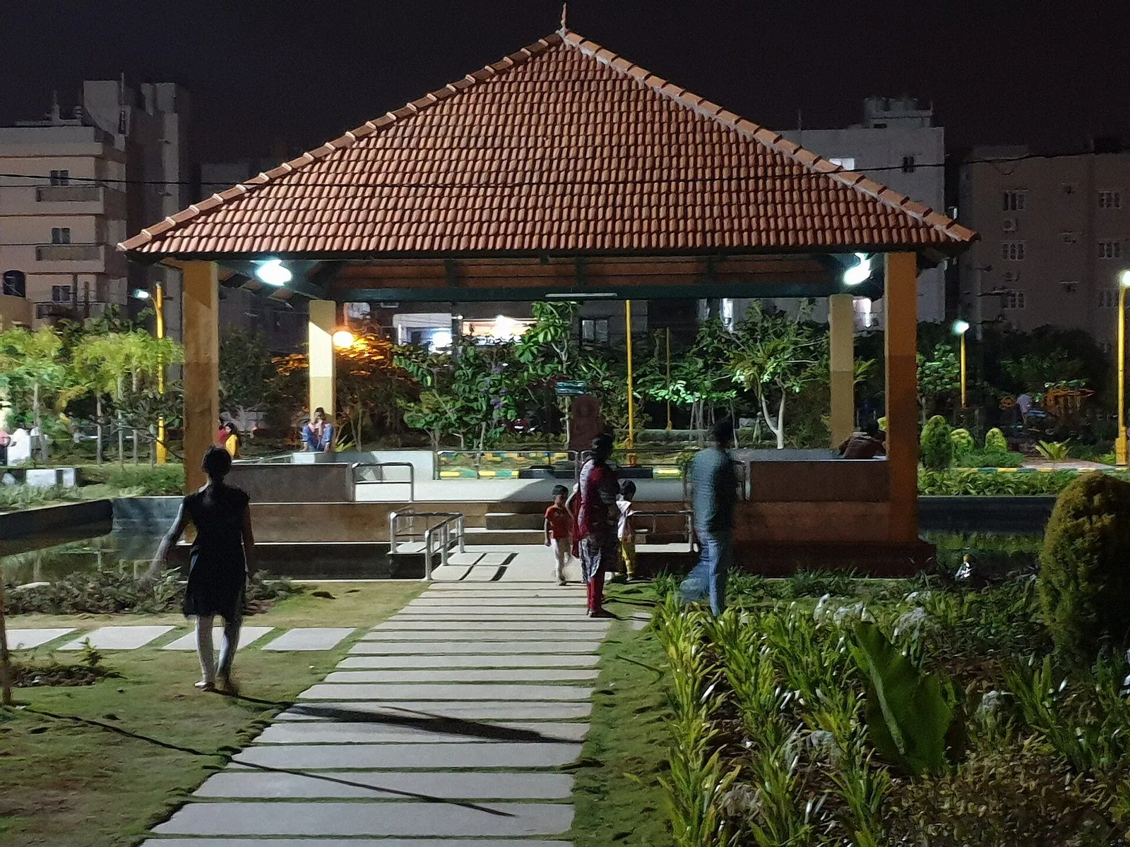 People walking on a stone path in a small park with a tiled pavilion, apartments behind