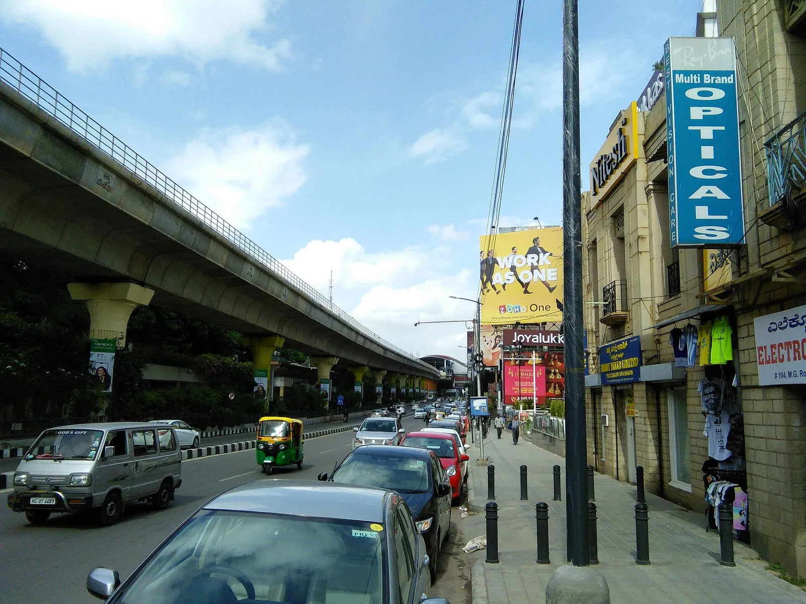 Cars and autos on MG Road beside shops, with the elevated metro line overhead